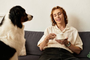 Genç bir adam kanepede dinleniyor, köpeği sabırla onu izlerken bir fincan çay içiyor..
