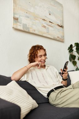 Gülümseyen genç adam evdeki kanepede dinleniyor, akıllı telefonuyla meşgul ve rahat hissediyor..