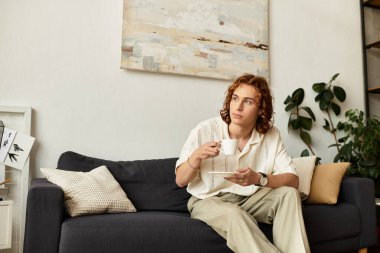 Genç bir adam rahat bir şekilde kanepede oturur, sıcak bir ev ortamında sıcak bir içkinin tadını çıkarır..