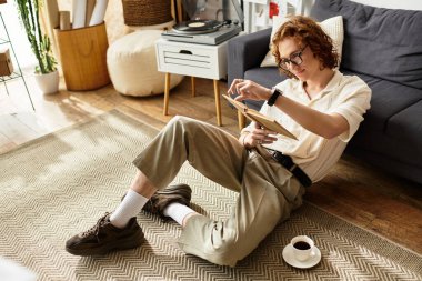 Kapalı mekanda, tableti ve kahvesiyle yerde dinlenen genç bir adamın görüntüsü..