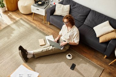 Genç bir adam rahat bir evde dizüstü bilgisayarıyla yerde rahatça oturuyor..