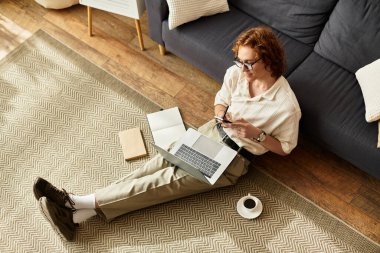 Konforlu bir oturma odasında, genç bir adam yerde oturur, bilgisayarına ve kahvesine odaklanır..