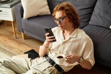Genç bir adam bir elinde bir fincan kahve, diğerinde akıllı bir telefonla yerde dinleniyor..