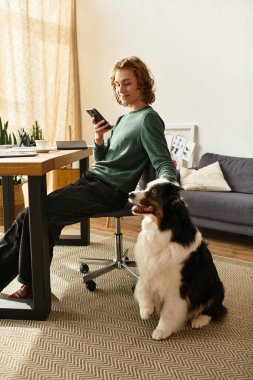 Döner sandalyede rahatlamış genç bir adam köpeğiyle telefonuna odaklanmış..