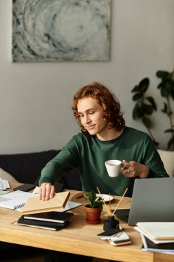 Genç bir adam evinde rahatça oturur, kahvesini yudumlar ve çalışma alanını odaklanarak düzenler..