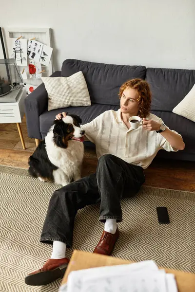 Genç bir adam köpeğiyle yerde dinleniyor, sıcak bir fincan kahvenin tadını çıkarıyor..