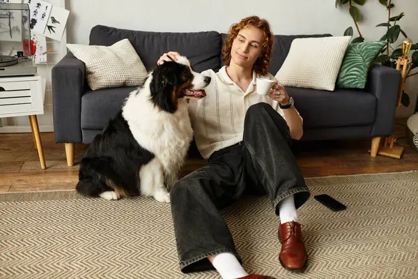 Genç adam kahvesini yudumlarken ve evde köpeğini okşarken rahatça yerde oturuyor..