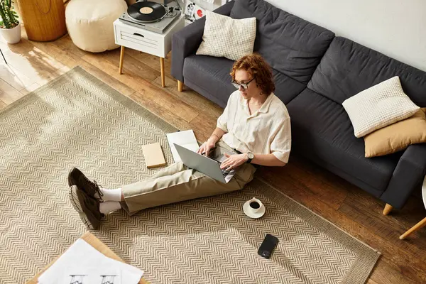 Genç bir adam rahat bir evde dizüstü bilgisayarıyla yerde rahatça oturuyor..