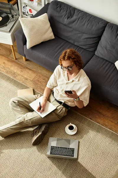 Genç adam elinde bir defterle yerde rahatça oturur, kahvesini yudumlar ve telefonunu kullanır..