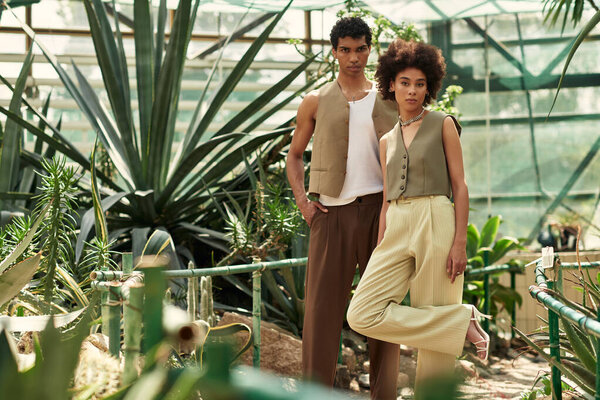 A couple stands closely together, surrounded by an abundance of plants in a bright greenhouse.