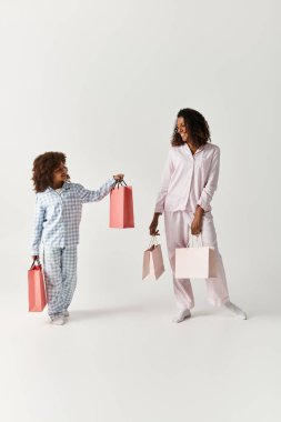 Siyah anne ve kız pijamalarıyla ellerinde pembe alışveriş çantaları, gri arka plan..