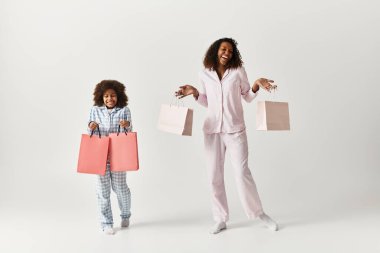 Bir anne ve kızı birbirlerine uygun pijamalarla alışveriş torbalarını tutarken neşeyle ışıldıyor..