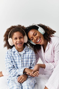 Siyah anne ve kızı pijamalarıyla gülüyor ve kulaklıkla müzik dinliyor, gri stüdyo.