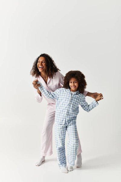 African American Mother and daughter wear pyjamas, holding hands, smiling, and dancing.