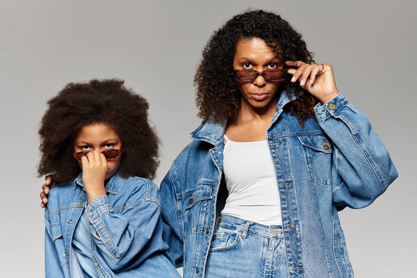 Black mom and daughter in matching denim, shades on grey.