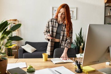 Kızıl saçlı güzel bir kadın, rahat bir çalışma alanında bitkilerle otururken telefonunu inceliyor.