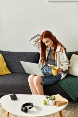 Genç ve güzel bir kadın kanepede oturup kahvesini yudumluyor ve laptopunu kullanıyor..