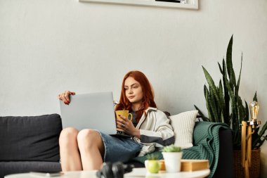 Rahat genç bir kadın kanepede oturur, sıcak bir içki yudumlar ve rahat bir yerde dizüstü bilgisayarını kullanır..