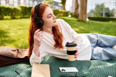 Genç ve güzel bir kadın yeşil bir battaniyeye uzanıyor, boş zamanlarının tadını kahve ve müzikle çıkarıyor..