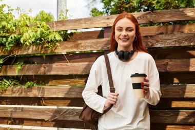 Parlak gülümseyen genç bir kadın elinde bir fincan kahve tutuyor, açık havada neşe saçıyor..