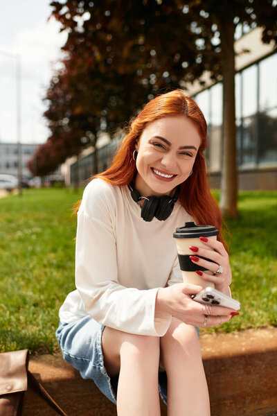 Beautiful young woman relaxes outdoors, sipping coffee and checking her phone while smiling.