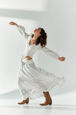 Genç bir kadın beyaz bir elbise ve kovboy çizmeleri içinde sevinçle kıvrılıyor, tarzını gösteriyor..