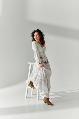 Kıvırcık saçlı genç bir kadın şık beyaz bir elbise içinde zarif bir şekilde poz verir..