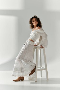 Beyazlar içinde şık bir genç kadın tabureye yaslanmış, şık kıyafetini hafif ışıkla renklendiriyor..