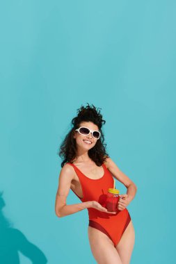 Kırmızı mayo giymiş şık genç bir kadın neşeli bir pozda içki tutuyor, kendine güveni tam..