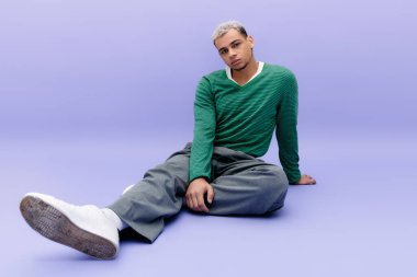 Saçları boyalı, kendine güvenen genç bir adam mor bir arka planda şık bir kıyafet içinde şık bir poz verir..