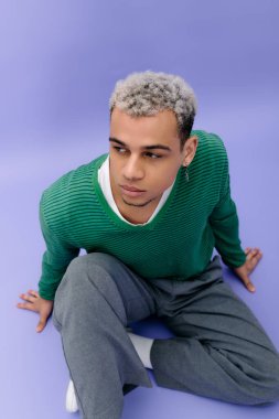 Saçları boyalı şık bir genç adam mor bir arkaplanda rahatça poz veriyor..