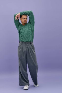 Boyalı saçlı genç bir siyah adam, tarzını modern bir kıyafetle mor bir arkaplana karşı sergiliyor..