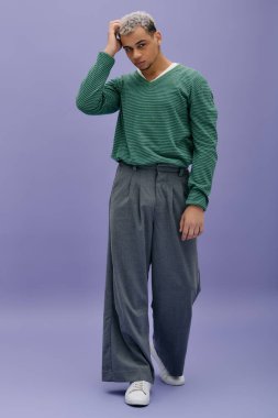 Saçları boyalı genç bir siyah adam mor bir arka planda kendine güvenen şık bir elbiseyle poz verir..