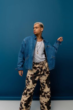Genç bir siyah adam şık bir kot ceketle parlak mavi bir fona karşı poz veriyor..