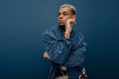 Kendine güvenen genç bir adam, mavi arka planda şık kot ceketini sergileyerek maharetli poz verir..