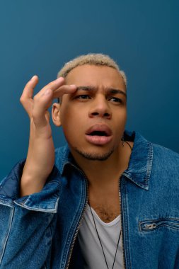 Boyalı saçlı genç siyah bir adam mavi bir arka fona karşı kot ceketiyle güvenle poz veriyor..