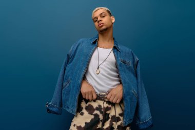 Şık boyalı saçları olan genç bir adam canlı maviye karşı kendine güvenen bir kot ceket giyiyor..