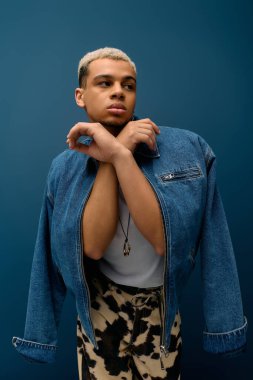 Genç siyahi bir adam, mavi bir arkaplana karşı, modaya uygun bir kot ceketle güvenle poz veriyor..