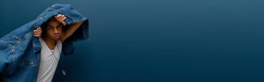 Tarz sahibi genç Afrikalı Amerikalı erkek, mavi arka planda kot ceket ve afiş giyerek rahatça poz veriyor.