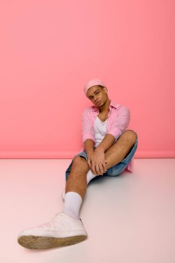 Genç bir siyah adam kendine güvenerek poz veriyor, canlı pembe bir arka plana karşı tarzını sergiliyor..