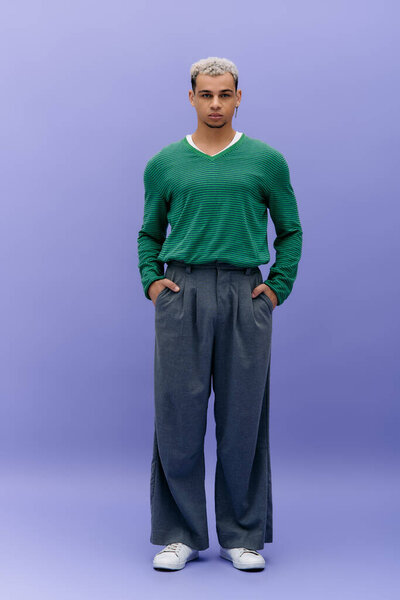 A young African American man showcases his trendy outfit while standing against a purple backdrop.