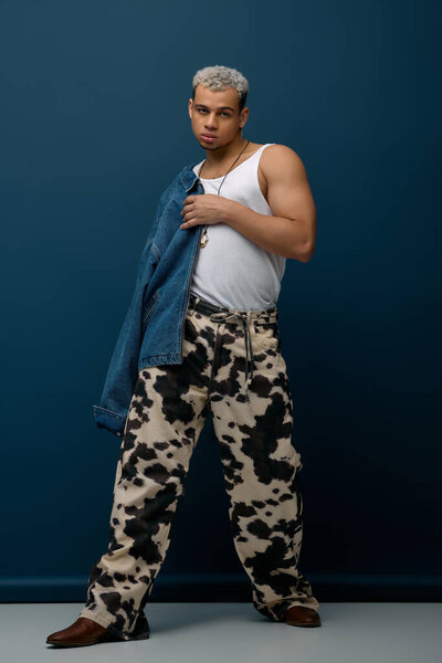 Fashion-forward young man in a denim jacket poses confidently against a striking blue background.