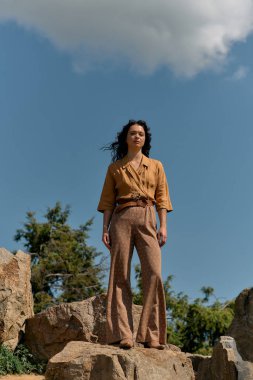 Kıvırcık saçlı kadın şık bir yaz kıyafetiyle kayaların üzerinde güven içinde poz veriyor..