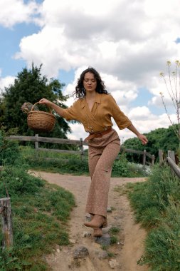 Curly-haired woman strolls along a scenic path while carrying a basket on a sunny summer day.