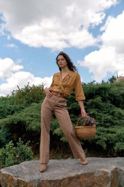 Genç bir kadın kendine güvenerek bir kayanın üzerinde duruyor ve şık yaz kıyafetiyle güneşin tadını çıkarıyor..