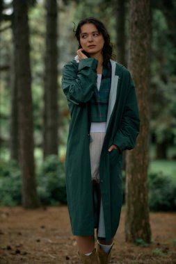 Bereketli bir yaz ormanında, kıvırcık saçlı genç bir kadın yansıtan şık bir yağmurluk giyer..