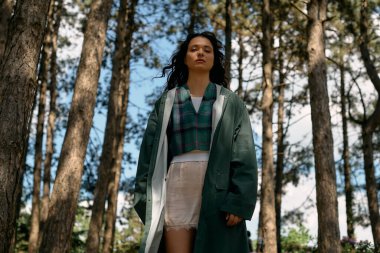 Yağmurluk giymiş genç bir kadın canlı yeşil ormanı keşfediyor, moda anlayışını gösteriyor..