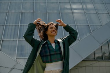 A cheerful young woman playfully poses outdoors in a trendy raincoat under a clear sky.