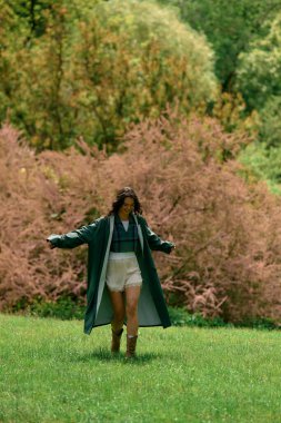 Young woman with curly hair twirls joyfully outdoors in a fashionable raincoat and shorts.