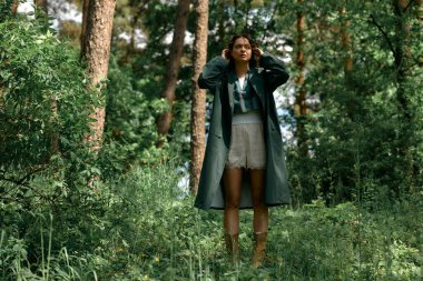 Kıvırcık saçlı kadın, dışarıda, ormanda şık bir yağmurlukla moda anlayışını sergiliyor..